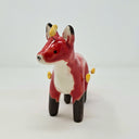 Ceramic sculpture of a red fox with a very fluffy tail, black paws, and has a simplistic drawn on face. Coming out from its body are small yellow flames.