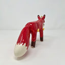 Ceramic sculpture of a red fox with a large fluffy tail and black paws. It holds a cap in its mouth, white and black with a Big Boss Robot logo on it. Small yellow flames come off of the fox's body.