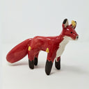Ceramic sculpture of a red fox with a very fluffy tail, black paws, and has a simplistic drawn on face. Coming out from its body are small yellow flames.