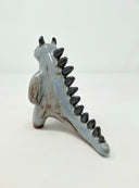 Ceramic sculpture of a bluish gray colored kaiju creature, standing on its hind legs with curved horns atop its head and small black spikes along its back.