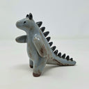 Ceramic sculpture of a bluish gray colored kaiju creature, standing on its hind legs with curved horns atop its head and small black spikes along its back.