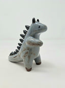Ceramic sculpture of a bluish gray colored kaiju creature, standing on its hind legs with curved horns atop its head and small black spikes along its back.