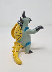 Ceramic sculpture of a kaiju style monster, standing on its hind legs with its arms situated out. It has a silver beak, mint colored body with a yellow belly, yellow fin on its back and a yellow dinosaur tail. It has silver spikes along its back and a red eye.
