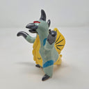 Ceramic sculpture of a kaiju style monster, standing on its hind legs with its arms situated out. It has a silver beak, mint colored body with a yellow belly, yellow fin on its back and a yellow dinosaur tail. It has silver spikes along its back and a red eye.