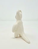 White glazed ceramic sculpture of a simplistic character standing on 2 legs, with its arms out at its side and 2 horns atop its head. Back view to show long tail.