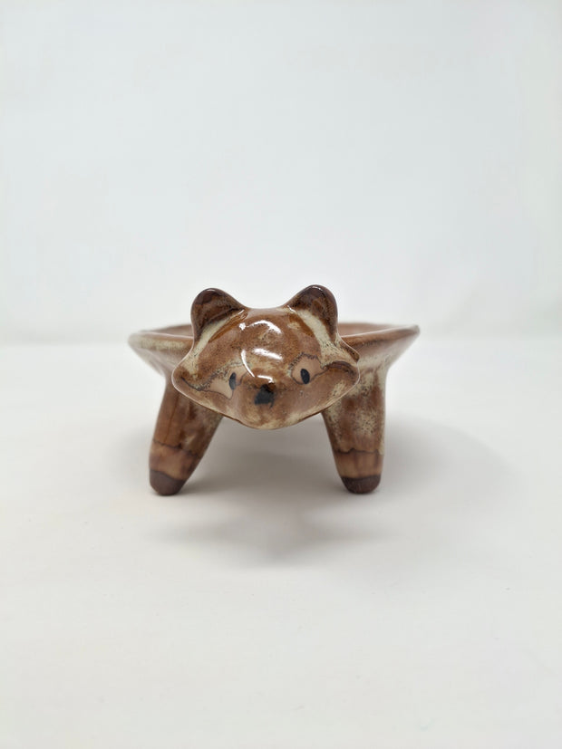 Ceramic plate or very shallow vessel, with a brown raccoon head in the front and 4 legs that act as stands for the vessel, attached. It has a fluffy, long tail.