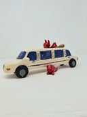 Ceramic sculpture of a white limo with small red devils hanging around it and inside of it, one pops out of the sun roof, the other hangs out and open door and the final is under the car.