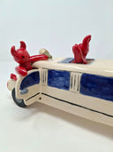 Ceramic sculpture of a white limo with small red devils hanging around it and inside of it, one pops out of the sun roof, the other hangs out and open door and the final is under the car.