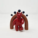 Glossy ceramic sculpture of a red devil with a simplistic, cute drawn on face. It stands in front of a brown miniature archway with blue ceramic spheres coming out of it from short wire posts.