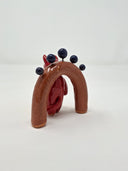 Glossy ceramic sculpture of a red devil with a simplistic, cute drawn on face. It stands in front of a brown miniature archway with blue ceramic spheres coming out of it from short wire posts.