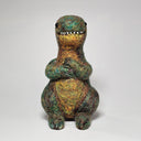 A handmade felted wool dinosaur toy with a textured, multi-colored body in shades of green, gold, and brown, standing against a neutral light gray background.