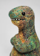 A handmade felted wool dinosaur toy with a textured, multi-colored body in shades of green, gold, and brown, standing against a neutral light gray background.