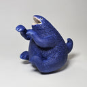 A blue textured dinosaur monster figurine with an open mouth showing white teeth, sitting on a light gray background.