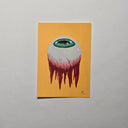 A vertical art print featuring a surreal illustration of a white eyeball with a green iris and dripping red blood, set against a solid yellow-orange background, displayed on a neutral light gray wall.