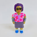 Clay sculpture of an older woman with greenish brown skin and short permed purple hair. She has gray wraps on both of her forearms, wears a purple and pink floral shirt and purple skirt.