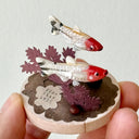 Small sculpture of 2 fish on a wooden base with paper plants and small rocks.