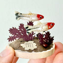 Small sculpture of 2 fish on a wooden base with paper plants and small rocks.