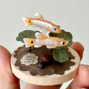 Small sculpture of 2 fish on a wooden base with paper plants and small rocks.