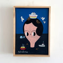 A framed folk art style painting of a large face with black hair and a striped sailor hat, with their body submerged in a body of water. Around them are small boats and birds, mounted on a white wall.