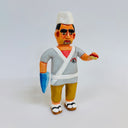Clay sculpture of a sushi chef, wearing a white sushi chef hat, a gray kimono shirt and holding a small piece of sushi in one hand. His other hand is a shark fin.