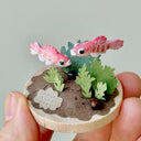 Miniature sculpture with two pink fish and green plants held by a hand against a light gray background.
