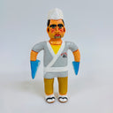 Clay sculpture of a sushi chef, wearing a white chef's hat, a gray kimono and sandals and socks. Both of his arms are shark fins.