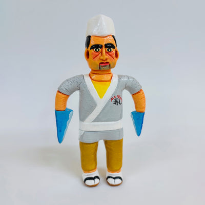 Clay sculpture of a sushi chef, wearing a white chef's hat, a gray kimono and sandals and socks. Both of his arms are shark fins.