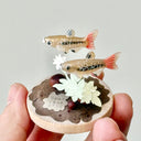 Miniature clay fish and plants on a circular base held by a hand against a light gray background