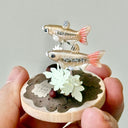 Small sculpture of 2 fish on a wooden base with paper plants and small rocks.