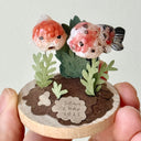 Small decorative piece with two goldfish and plants on a circular base, held by a hand against a light background.