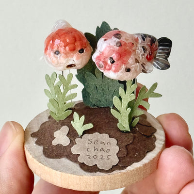 Small decorative piece with two goldfish and plants on a circular base, held by a hand against a light background.