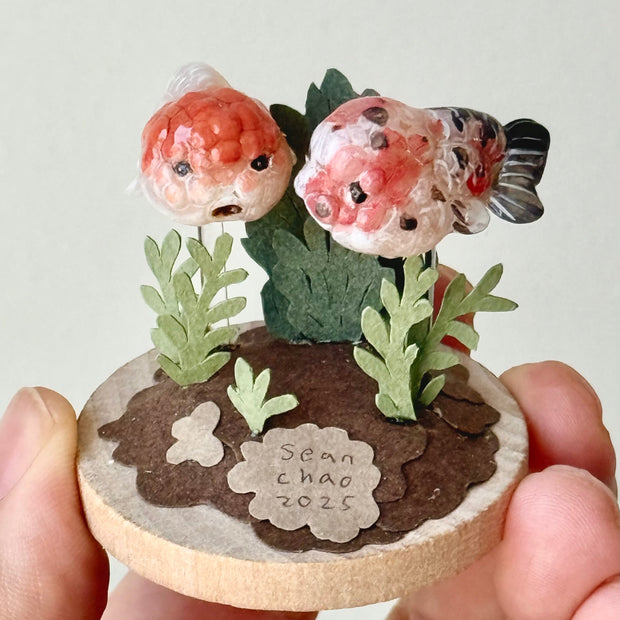Small decorative piece with two goldfish and plants on a circular base, held by a hand against a light background.
