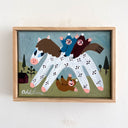 A framed folk art style painting featuring a stylized white horse with black spots ridden by two children in blue and brown outfits. Below the horse is another child, wearing a light brown outfit that has fallen off.