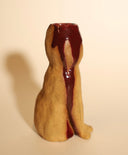 Ceramic sculpture of a tall brown cat, sitting and looking forward with simplistic eyes and a nose. Its head is cut open in a cross section, with blood spilling out.