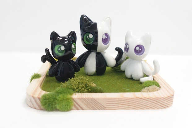 Three cat shaped figurines, with cute, large anime style eyes standing on a wooden tray with fake grass. They all have 2 tails.