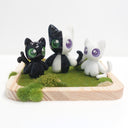 Three cat shaped figurines, with cute, large anime style eyes standing on a wooden tray with fake grass. They all have 2 tails.