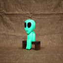 Whittled wooden sculpture of a green alien with a large head and black eyes. It has a slight frown and sits on a bench. It glows under blacklight.