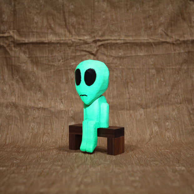 Whittled wooden sculpture of a green alien with a large head and black eyes. It has a slight frown and sits on a bench. It glows under blacklight.