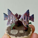Small sculpture of 2 purple striped angel fish, facing one another on a wooden base with stones and paper plants.