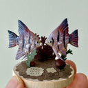 Small sculpture of 2 purple striped angel fish, facing one another on a wooden base with stones and paper plants.