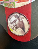 Sculpture of a doll with a wide head, closed eyes and its lips pursed in a kiss. It has large ears and wears a red robot helmet and has a red robot body with the limbs dangling. Its attached to a plaque with a collage of Giant Robot magazine covers.