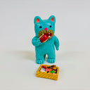 Clay sculpture of a small blue cat, standing on 2 legs like a person, eating a piece of sushi. It stands in front of a sushi bento box on the ground.