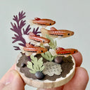 Small sculpture with clay fish and plants held by a hand against a light gray background