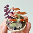 Small sculpture with clay fish and plants held by a hand against a light gray background