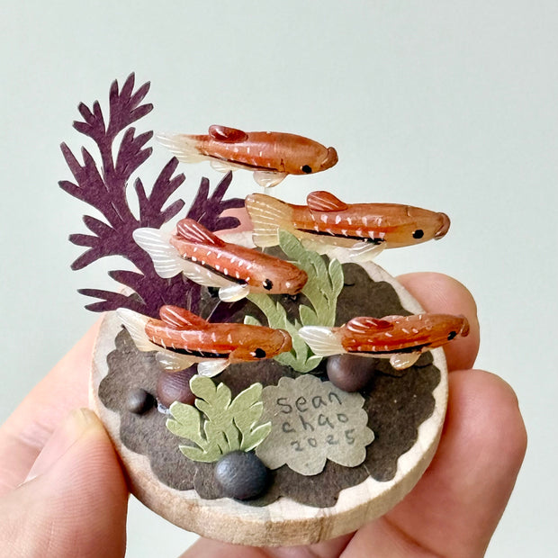 Small sculpture with clay fish and plants held by a hand against a light gray background