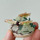 Small sculpture with clay fish and plants held by a hand against a light gray background