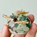 Small sculpture with clay fish and plants held by a hand against a light gray background