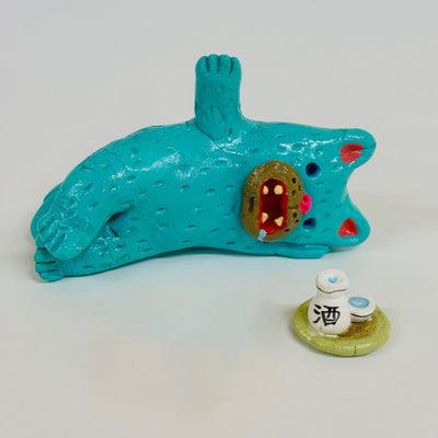 Blue clay cat laying next to a tray with sake on it.