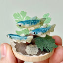 Small sculpture with clay fish and plants held by a hand against a light gray background