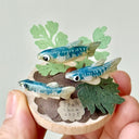 Small sculpture with clay fish and plants held by a hand against a light gray background